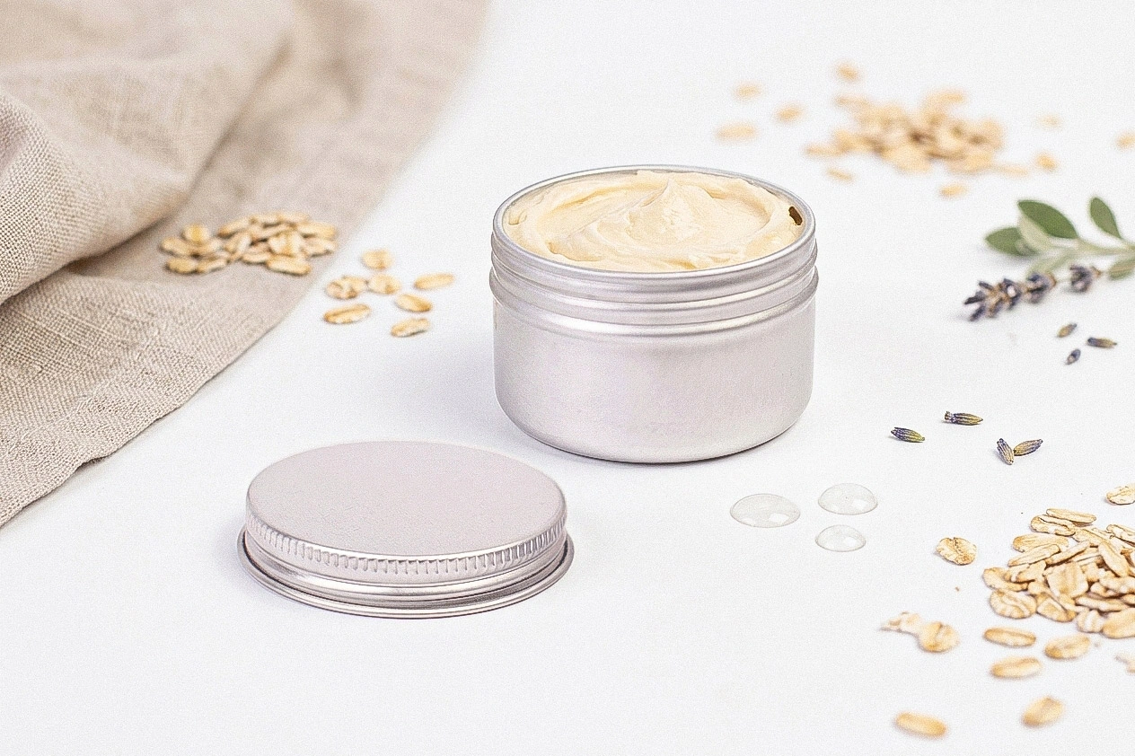Offene, silberfarbene Aluminiumdose mit Schraubdeckel, gefüllt mit einer hellen Creme, auf weißem Untergrund mit Haferflocken, Lavendel und grünen Blättern dekoriert