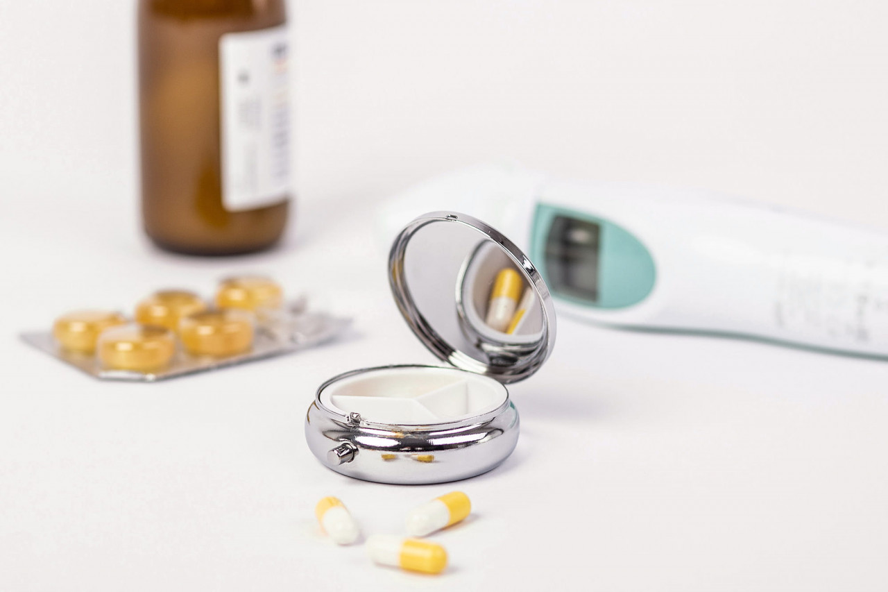 Geöffnete runde Pillendose aus silberfarbenem Metall mit drei Innenfächern und integriertem Spiegel, fotografiert mit Tabletten und Medizin im Hintergrund.