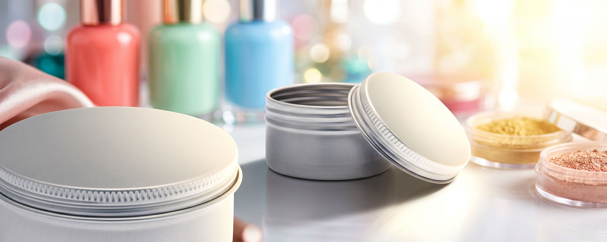 Offene silberne Metalldosen mit Kosmetikpuder und Beauty-Produkten auf weißer Oberfläche; elegante Produktaufnahme mit pastellfarbenen Nagellacken im Hintergrund.